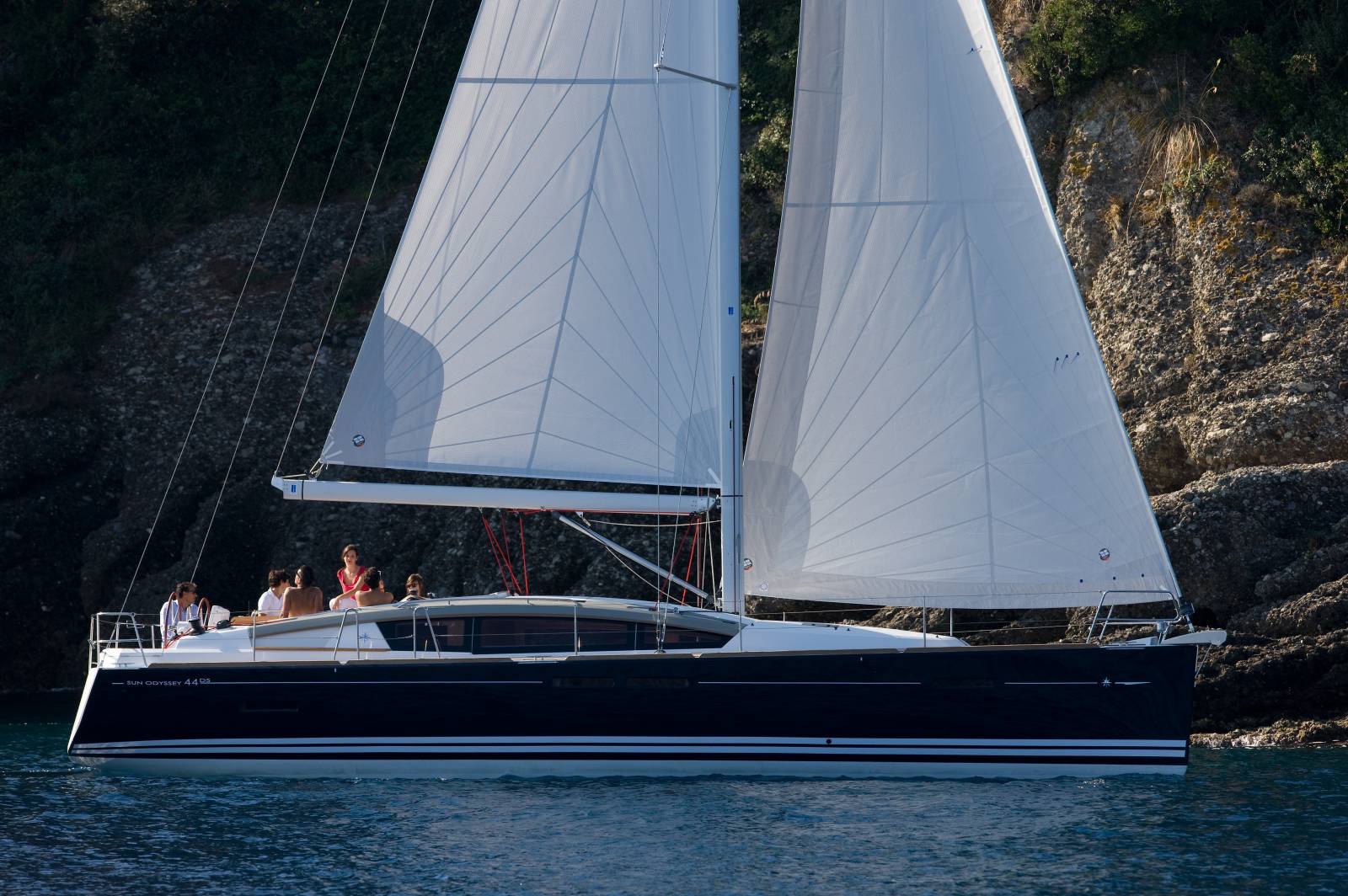 Jeanneau Sun Odyssey 44 DS under full sail, navy hull, with the rocky coast in the background